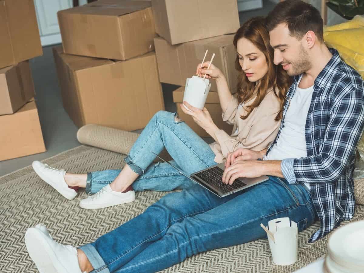 a man and woman surrounded by moving boxes eating take out and looking at a computer