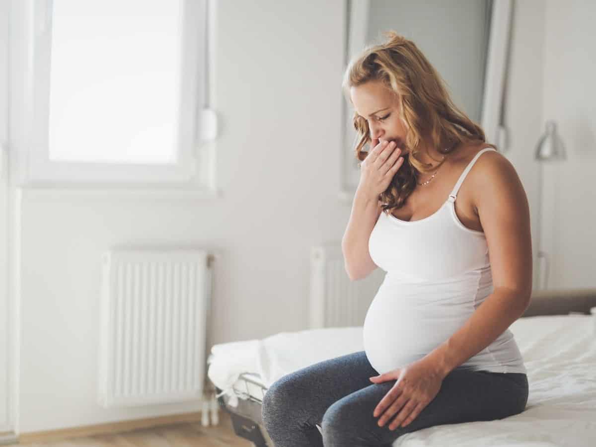 a pregnant woman who looks nauseous while sitting on her bed.