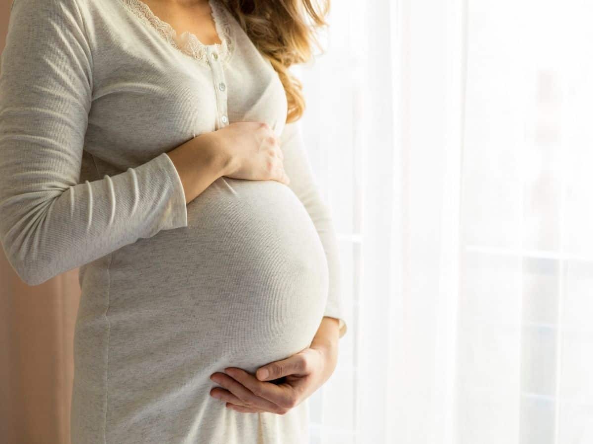 a pregnant woman standing by a window with her hands on her growing belly