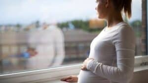 a pregnant woman looking out a window with a hand on her baby bump