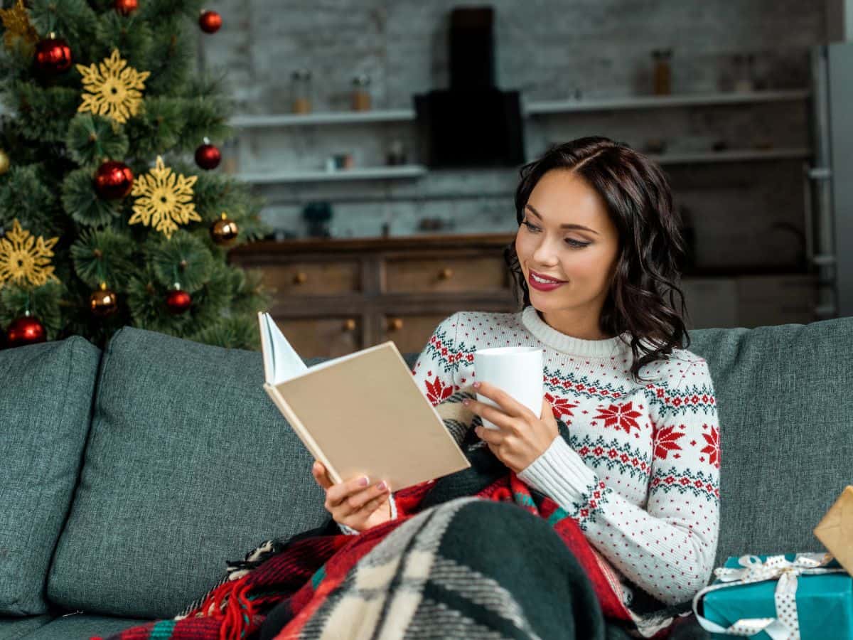 a woman sitting on a couch reading with a Christmas tree in the background and presents beside her.
