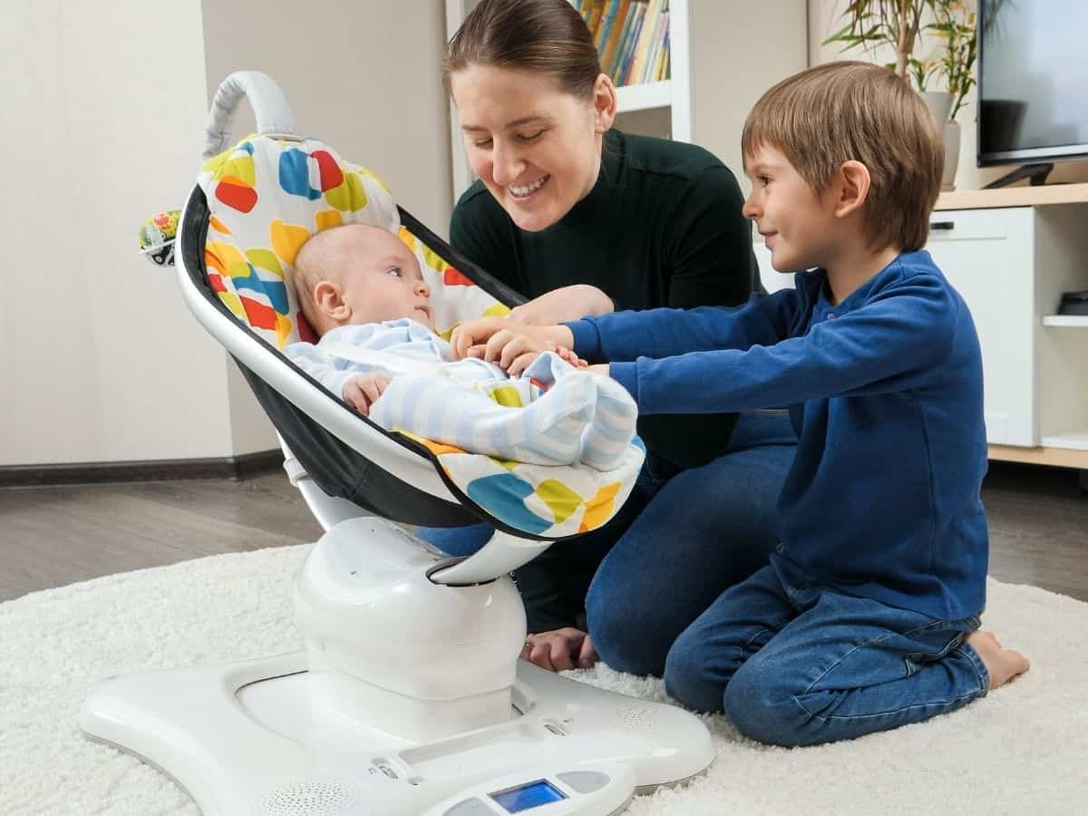 an infant in a colorful baby swing with his mom and big brother watching him