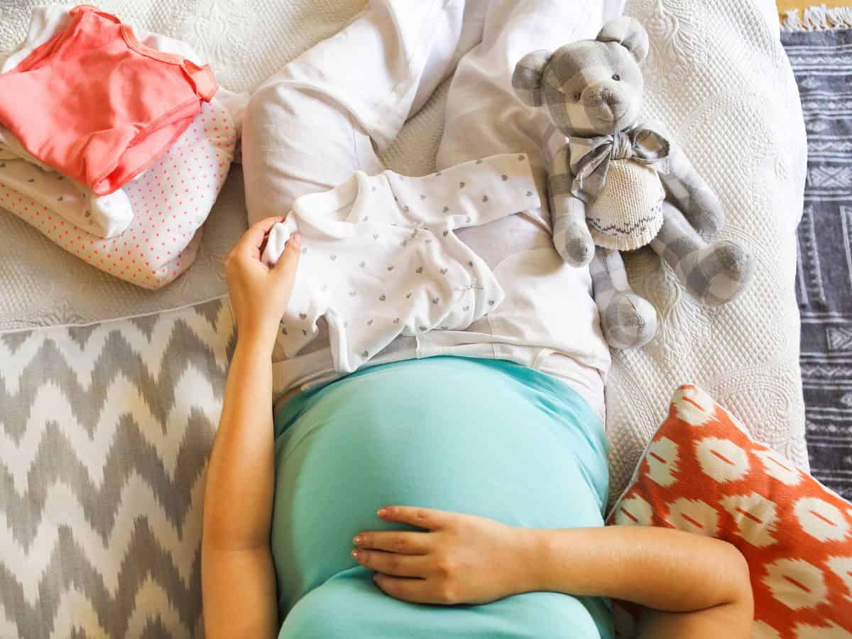 a pregnant woman folding baby clothes on her bed
